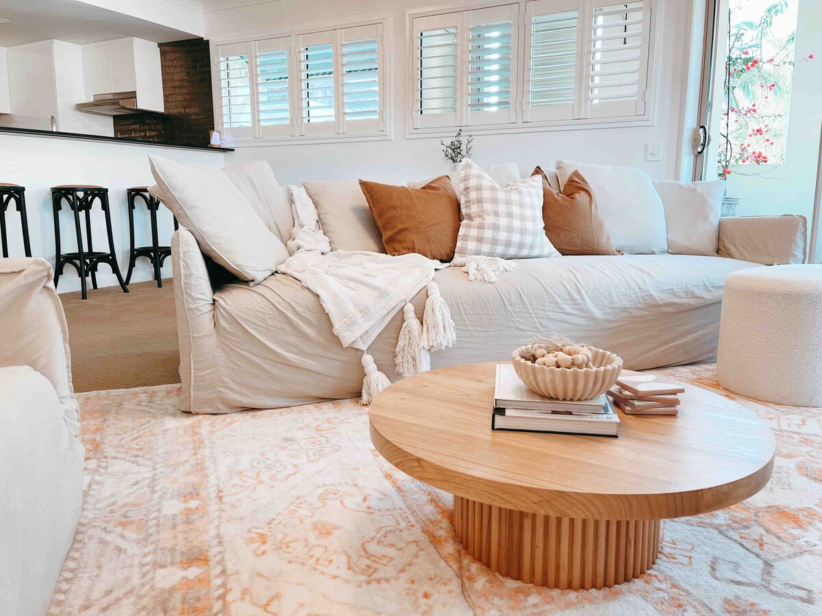 A spacious living area features a light-colored sofa adorned with brown and white decorative pillows. A soft area rug lies underneath a round wooden coffee table, which is topped with a bowl of decorative items and books. Natural light fills the room through shuttered windows.