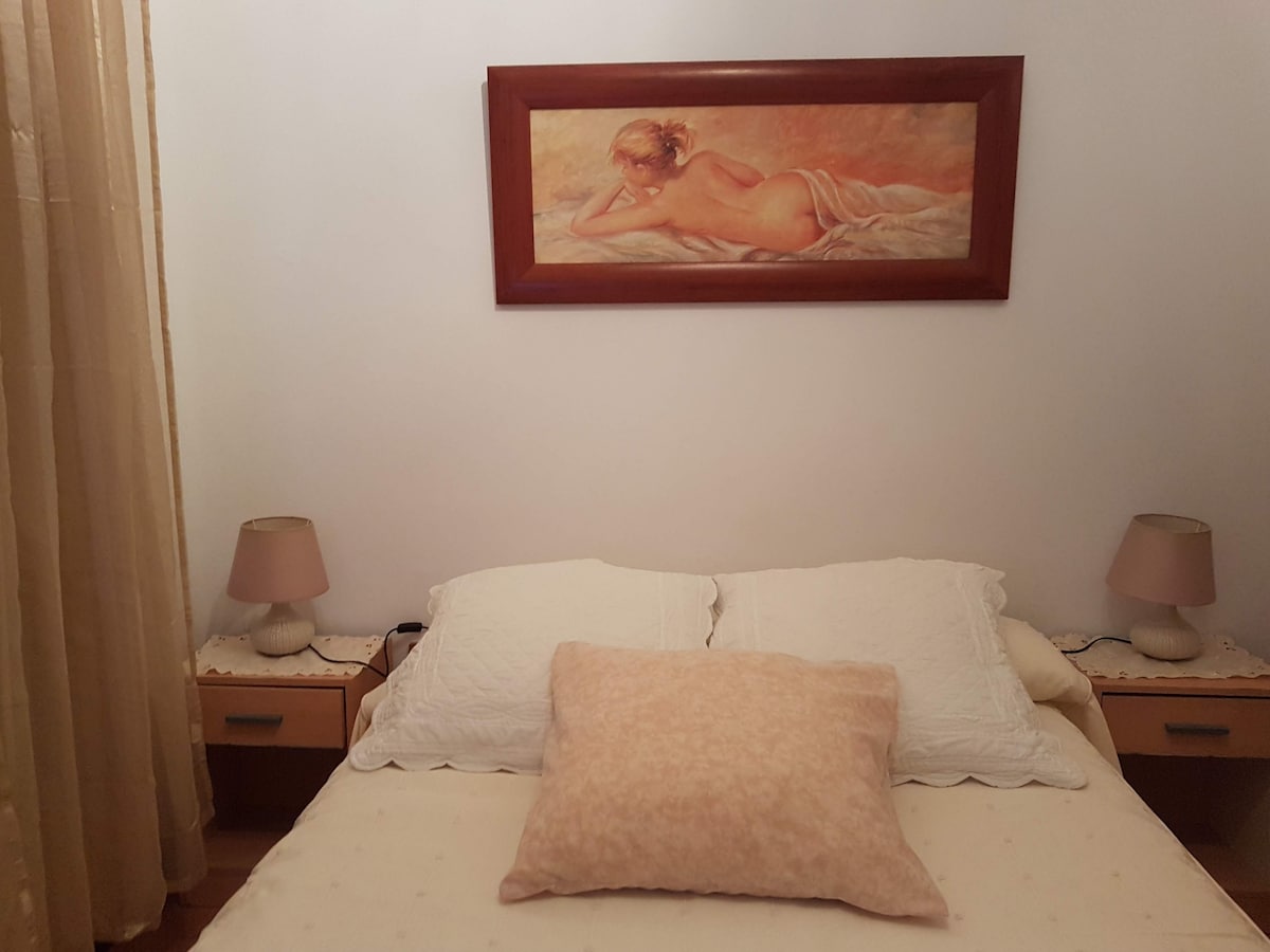 The spacious bed is adorned with a soft blanket and a large pillow. Two bedside tables with lamps are positioned on either side. A framed painting is hung on the wall above the bed, while soft curtains allow natural light to filter into the room.