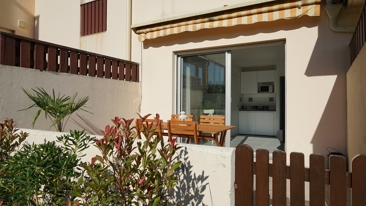 A terrace featuring a wooden dining table with chairs is visible, surrounded by low-maintenance greenery. Sunlight illuminates the space, and a striped awning provides shade. Sliding glass doors leading into the interior can be seen, allowing a glimpse of the kitchen area.