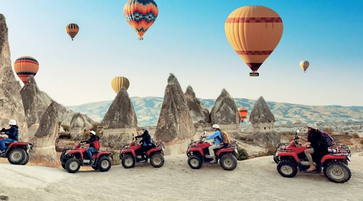 Several individuals are riding all-terrain vehicles on a dusty trail, surrounded by unique rock formations. Colorful hot air balloons float gently above, creating a vibrant contrast against the clear blue sky.