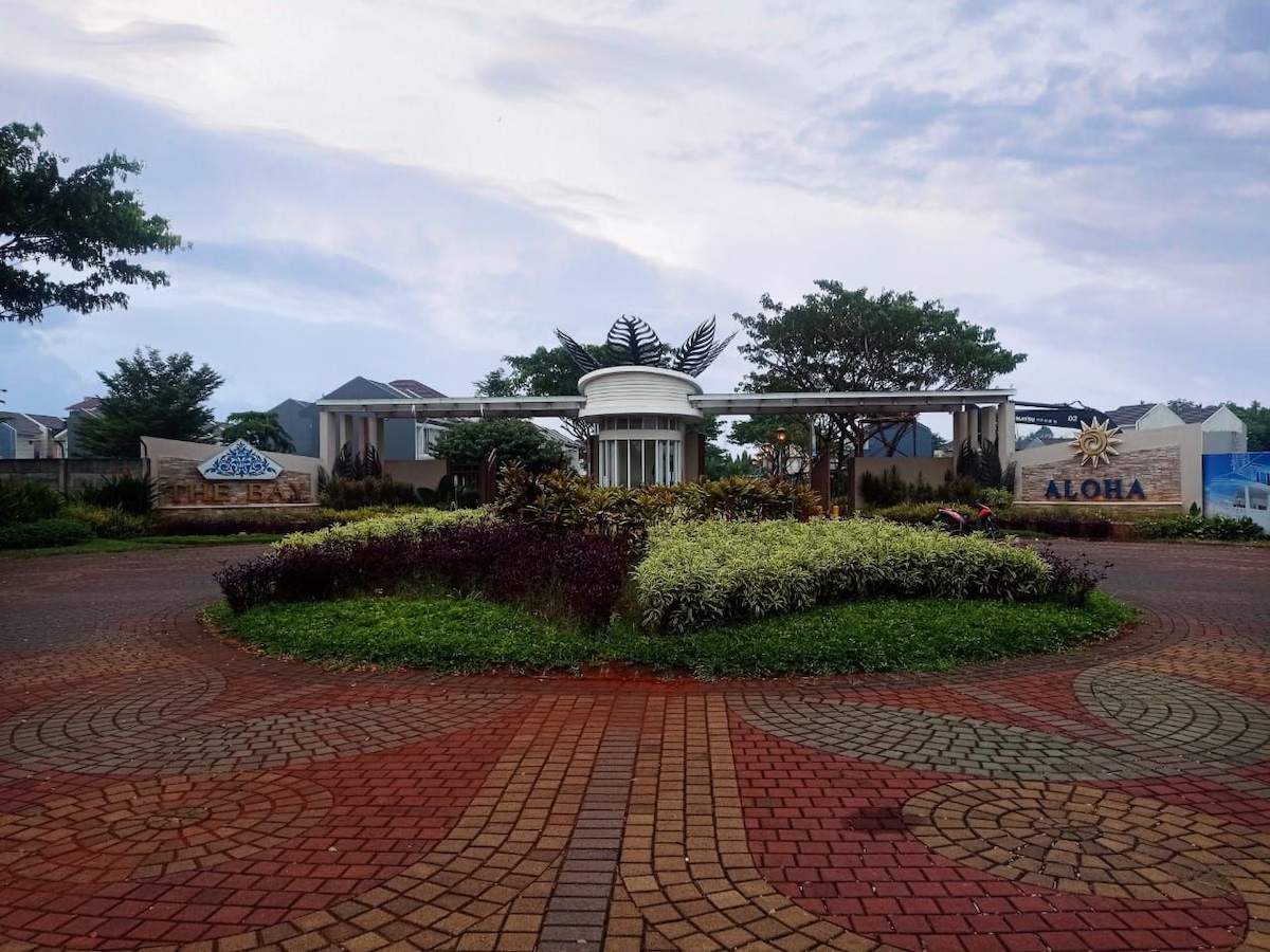 The entrance to Paradise Resort is showcased, featuring a circular driveway surrounded by manicured landscaping. Natural foliage and colorful flowers complement the decorative elements at the entrance, with a prominent sign that reads 'ALOHA' welcoming guests to the community.
