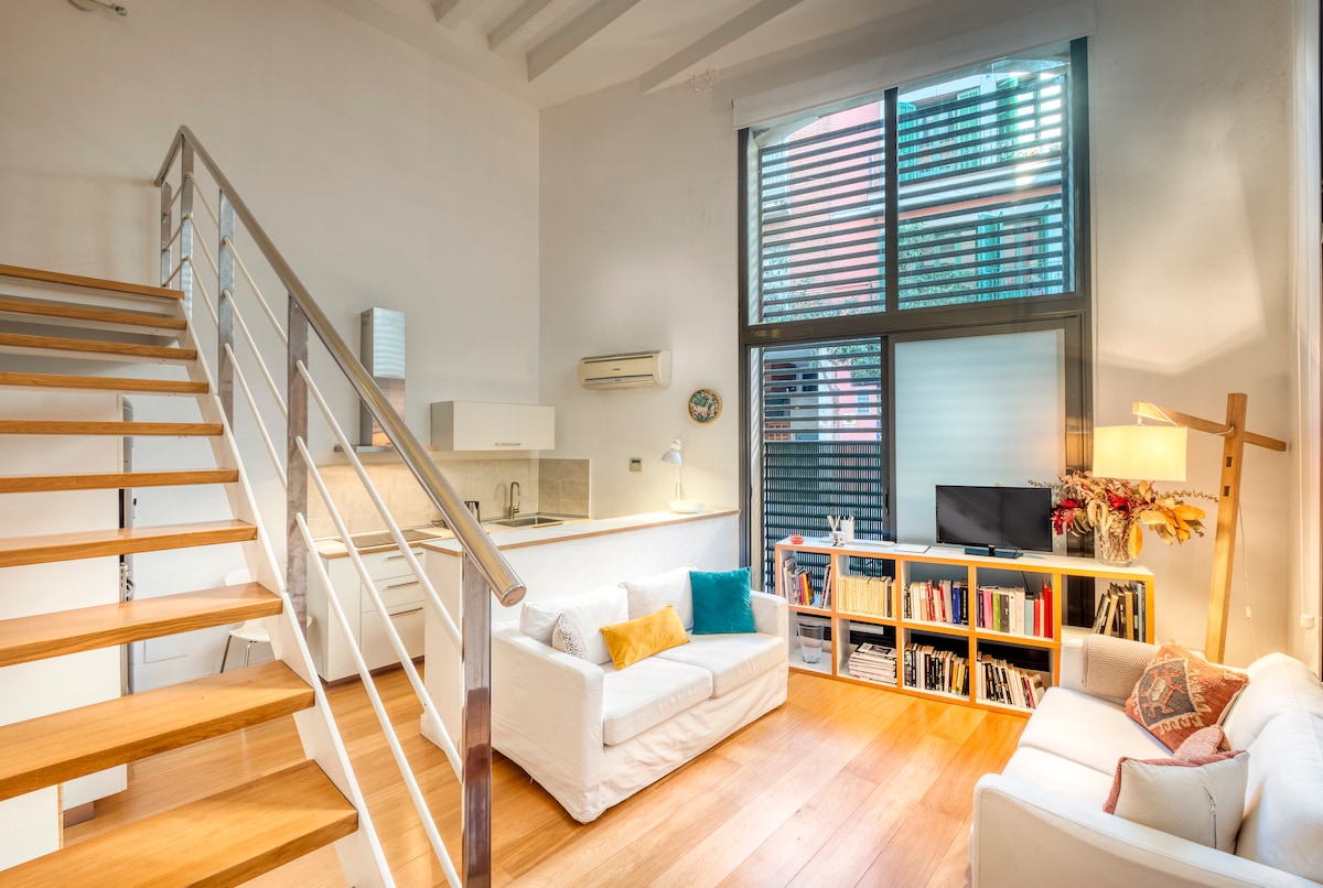 A bright living area features large windows with horizontal slats. A staircase leads to an upper level. Two white sofas with decorative pillows are positioned around a modern coffee table, while a collection of books is displayed on a nearby shelf.