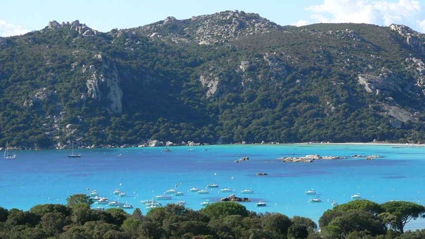A scenic view of a tranquil bay is presented, showcasing clear turquoise waters dotted with boats. Lush green hills rise in the background, complemented by rocky outcrops. A harmonious blend of natural elements creates a serene coastal landscape.