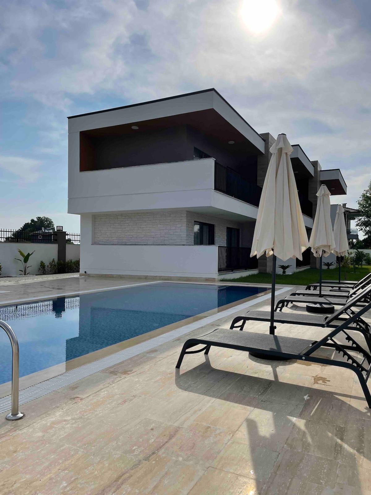 A modern building with a clean design features spacious balconies overlooking a tranquil pool area. Lounge chairs are positioned beside the pool, accompanied by standing umbrellas. The clear sky above is complemented by sunlight reflecting off the water's surface.