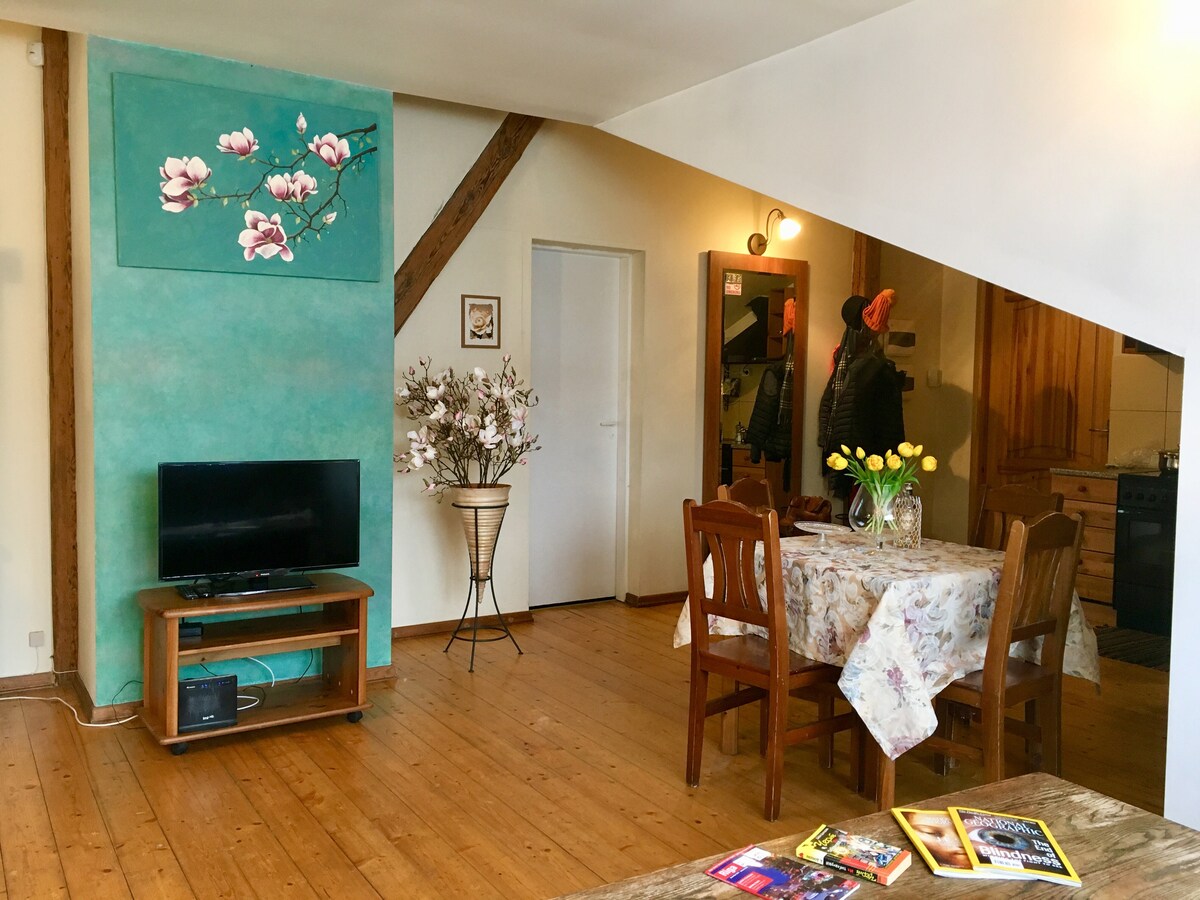 A spacious living area is furnished with a wooden dining table and chairs, accompanied by a television on a stand. A decorative vase with flowers enhances the ambiance. A vibrant wall art piece featuring magnolias adds a touch of color to the light-filled room.