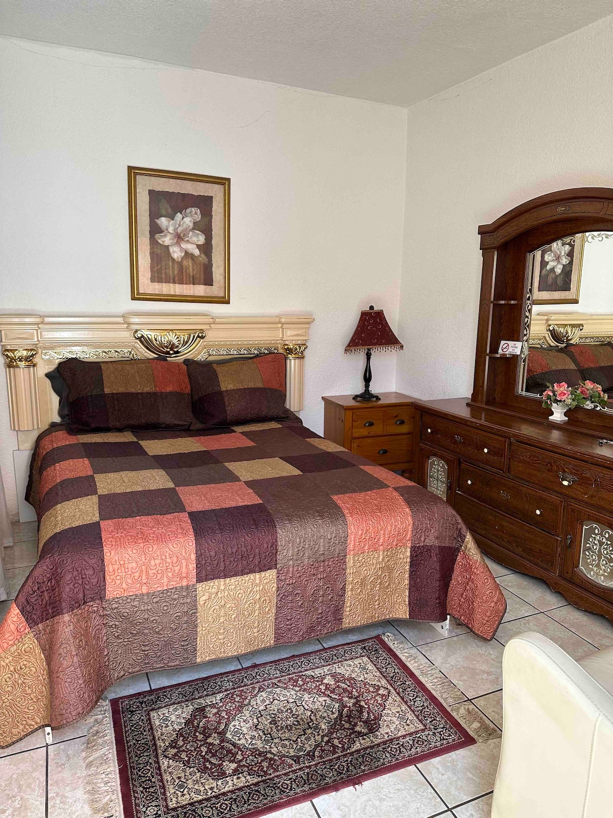 A cozy bedroom features a quilted bedspread in rich earth tones, complemented by patterned wall decor. A wooden dresser with a mirror is present, along with a bedside table and a lamp, creating a functional and inviting space.