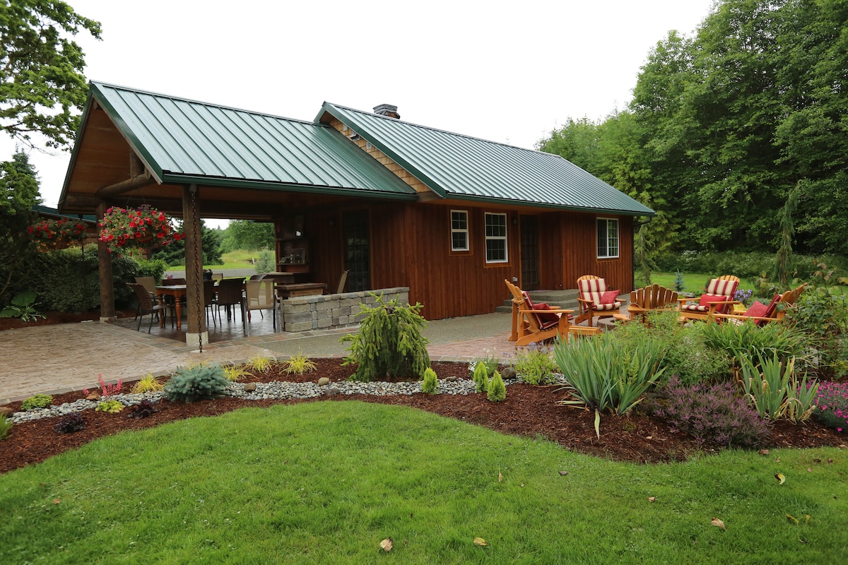 A charming cabin features a green metal roof and wooden exterior, set against a lush backdrop. A spacious patio area is accompanied by outdoor seating, with vibrant flowers lining the garden beds, creating a serene outdoor space.