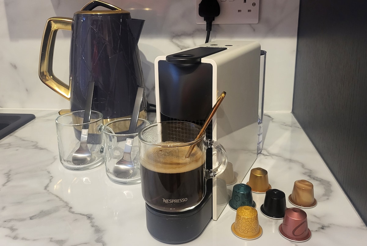 A modern coffee station is presented, featuring a Nespresso machine accompanied by a glass cup filled with freshly brewed coffee. Various coffee capsules are arranged nearby, and clear glass cups sit on the marble countertop, enhancing the contemporary kitchen aesthetic.