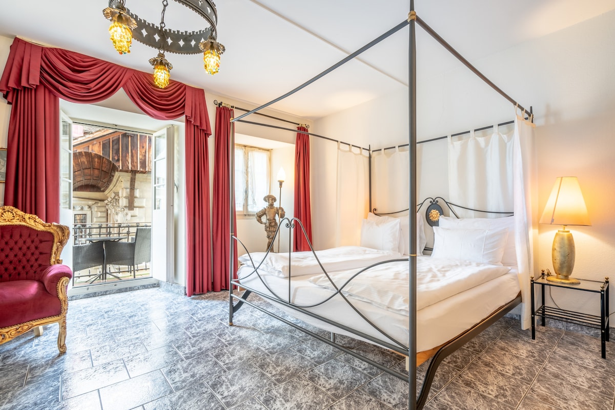 A spacious room features a four-poster bed with elegant drapes. An inviting armchair adds a touch of comfort next to the window, which opens to a balcony. Soft lighting is provided by a stylish lamp and a decorative chandelier illuminates the space.