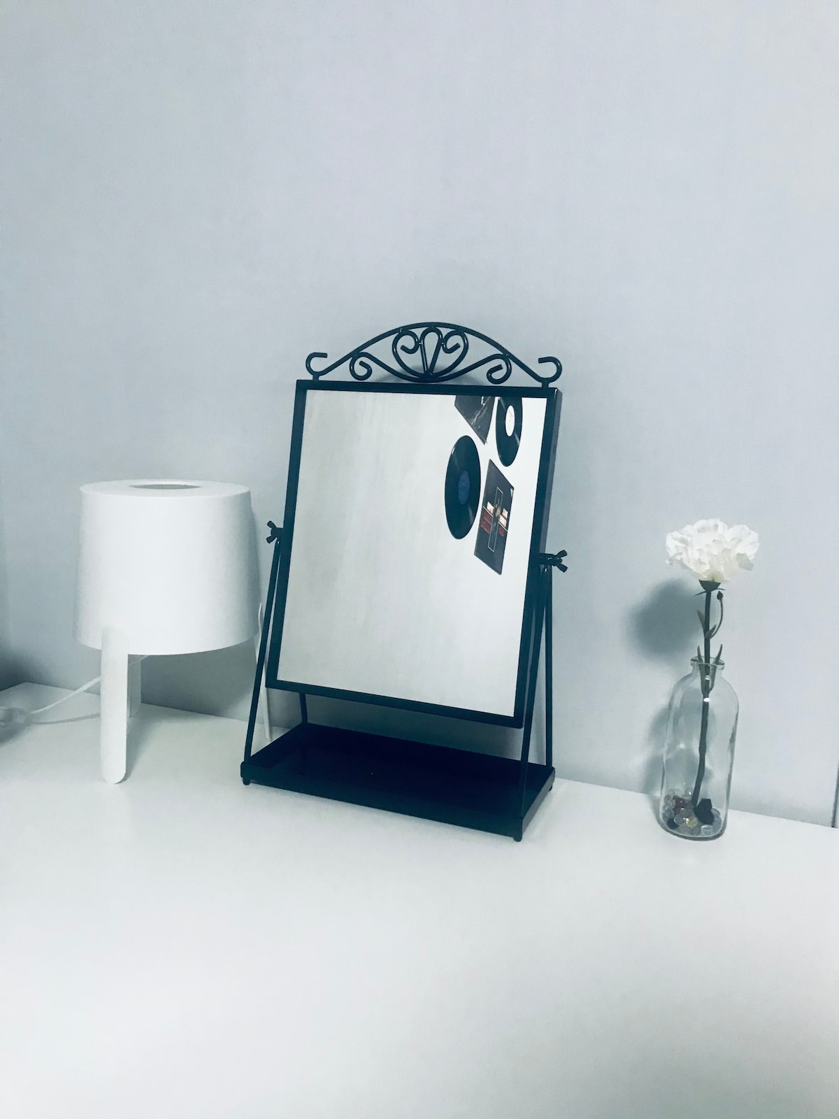 A minimalistic dressing table is featured with a tabletop mirror supported by an ornate black frame. A simple white lamp and a slender vase with a flower are positioned nearby, creating a serene and functional workspace.
