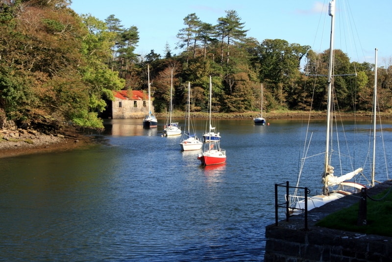 A serene waterway is framed by trees, with boats gently bobbing in the calm waters. A small building with a red roof sits on the shore, surrounded by lush greenery. The scene captures a peaceful moment in nature.