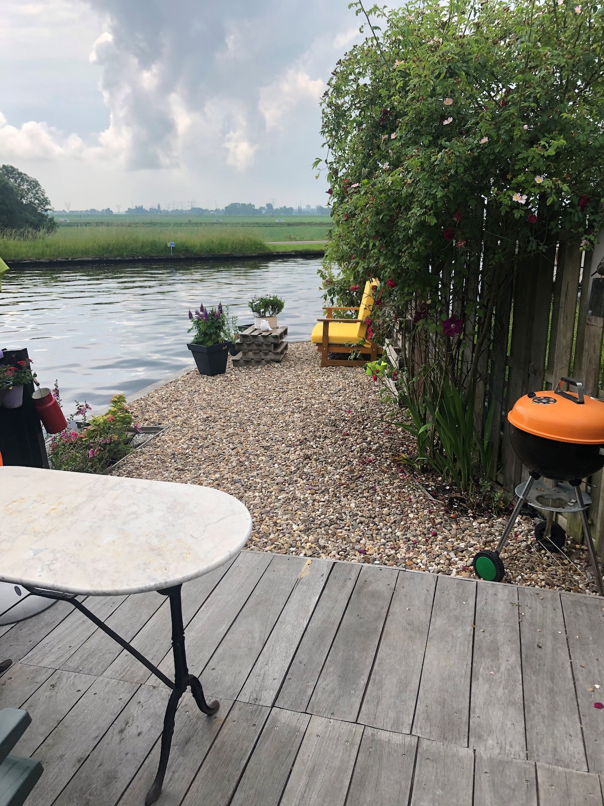 A private garden area features a gravel path leading to the water's edge. A round marble table and chairs are positioned nearby, along with colorful potted plants. A vibrant orange grill and a lounge chair add comfort to the serene outdoor space, surrounded by green scenery.