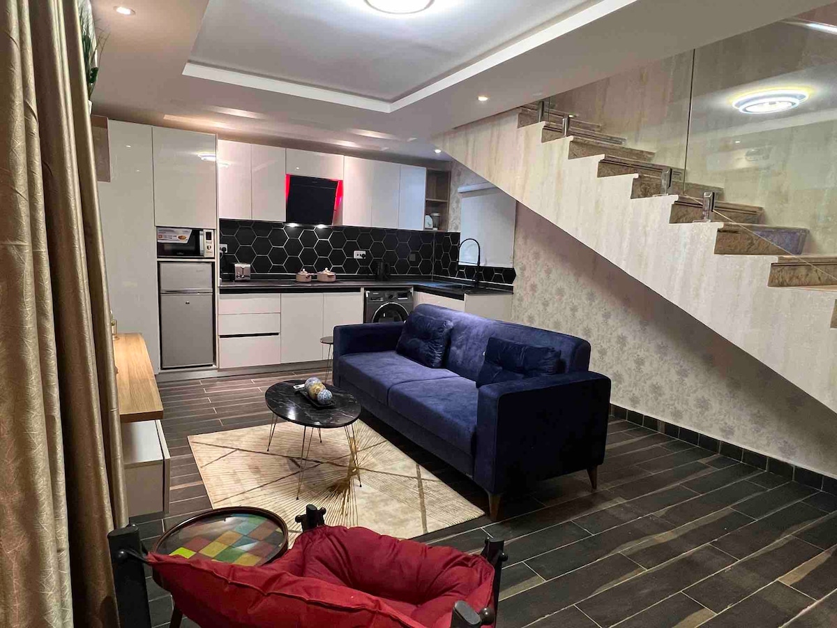 The living room features a modern blue sofa facing a sleek coffee table, set against a backdrop of stylish kitchen cabinetry. An open staircase leads to the upper level. Soft lighting enhances the contemporary design, while a patterned rug adds warmth to the space.