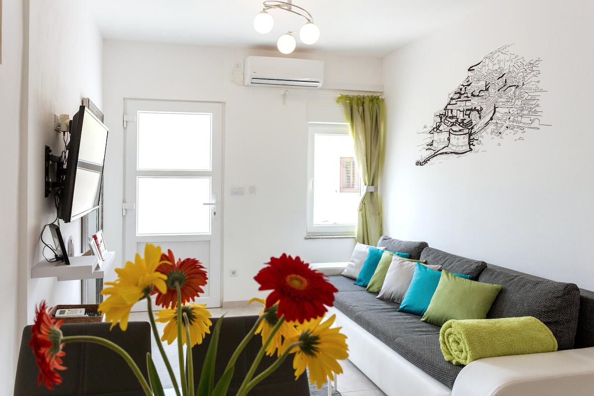 A light-filled living area is showcased, featuring a gray sofa adorned with colorful pillows. A wall-mounted TV is visible next to a window dressed with sheer curtains. A vibrant floral arrangement in the foreground adds a touch of color to the space.