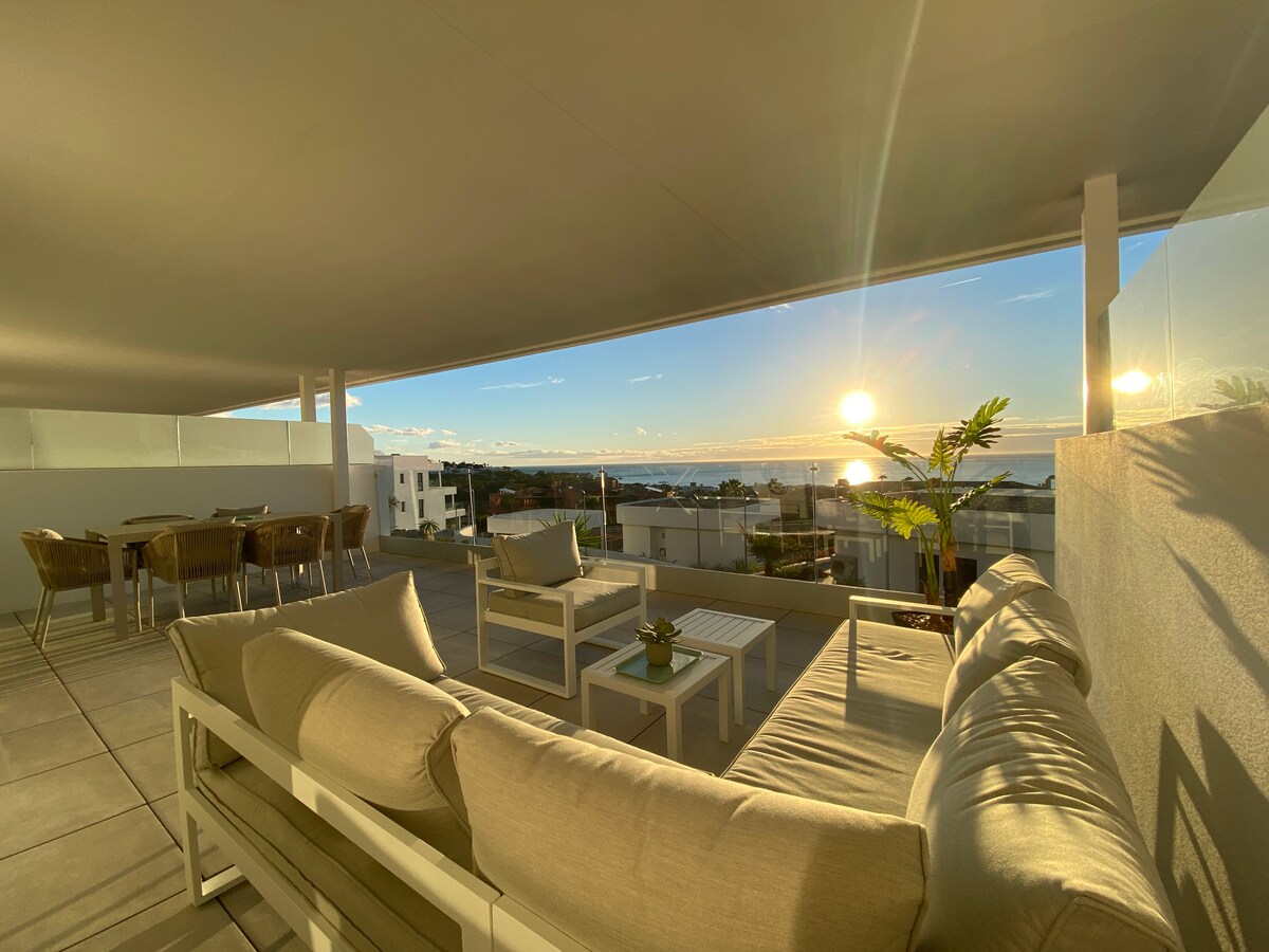 An expansive outdoor terrace is presented, featuring a comfortable seating area with light-colored cushions. A dining set for six is visible, complemented by a backdrop of the sea and a sunset sky, enhancing the serene environment.