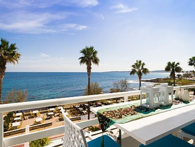 A balcony scene features a white table set for outdoor dining, with decorative lanterns and a vibrant green runner. Palm trees are visible in the foreground, while the tranquil sea extends into the horizon under a clear blue sky.
