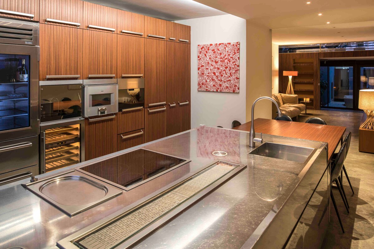 A modern kitchen is displayed with sleek wooden cabinetry and high-end appliances. A spacious countertop features a built-in stove and sink, complemented by an artistic wall piece. Soft lighting enhances the contemporary design and creates a functional cooking space.