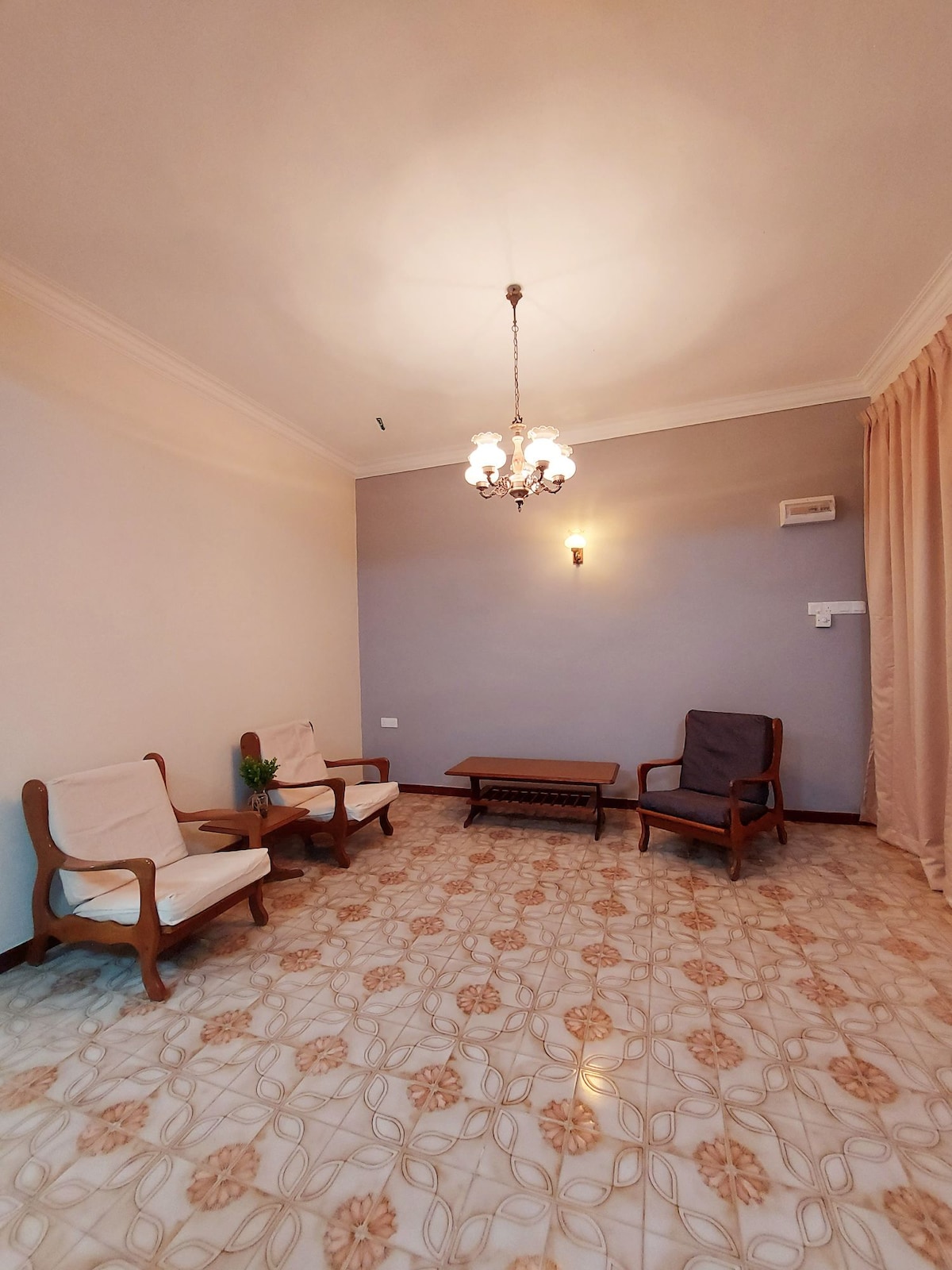 A spacious living area is presented with two wooden chairs and a small coffee table. The tile floor features a floral pattern. Soft lighting is provided by a chandelier and wall-mounted fixtures. Light curtains frame a window, adding to the bright ambiance.