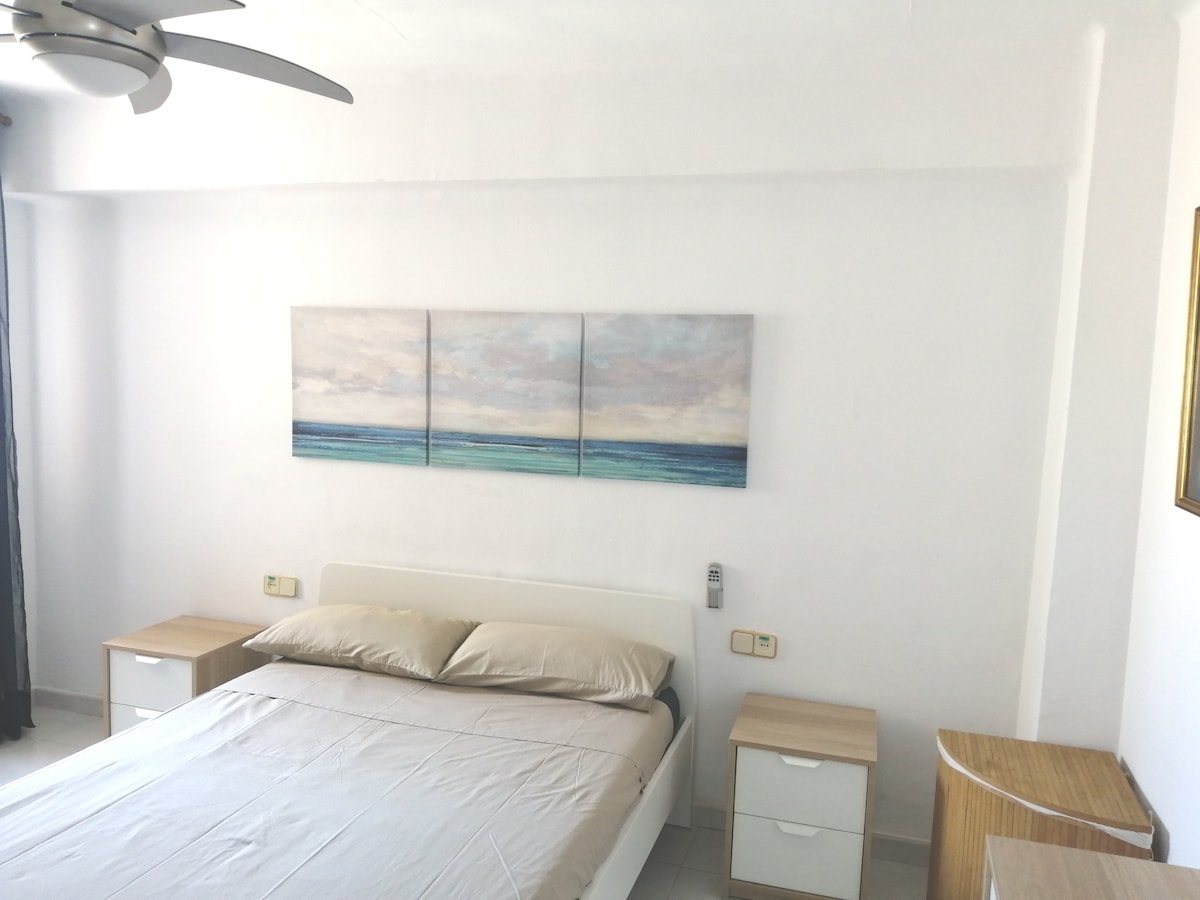 A spacious bedroom is presented with a large bed featuring a neutral-colored duvet. Two bedside tables with lamps are positioned on either side. Above the bed, a triptych artwork depicting a calm seascape adds color to the white walls, complemented by natural light from the window.