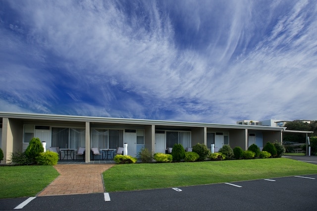 A low-rise building is shown with multiple units, each featuring a small patio area. Lush green shrubs and well-kept grass enhance the exterior, while expansive blue skies filled with soft white clouds create a serene backdrop.