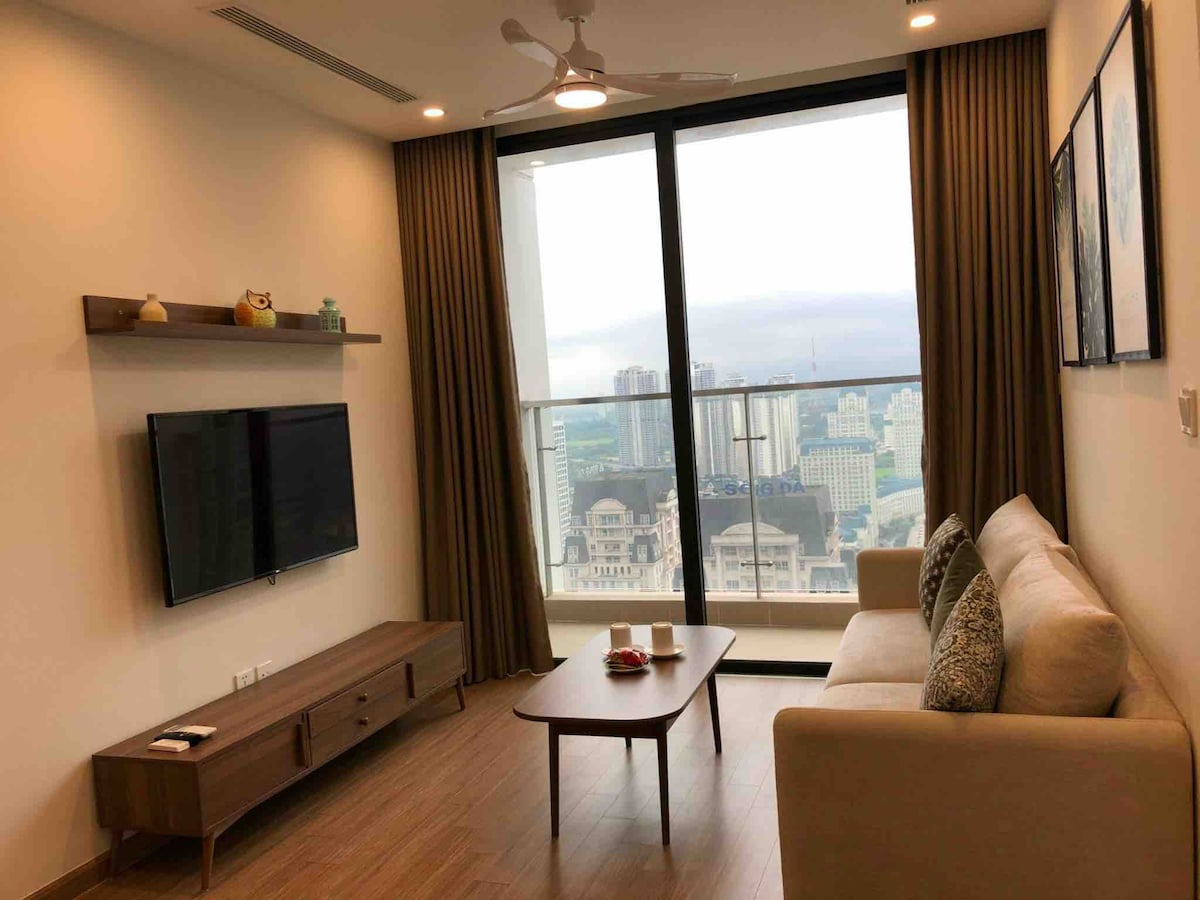 A modern living space features a spacious sofa positioned across from a wall-mounted television. A wooden coffee table sits in front of the sofa, and large windows provide a view of the cityscape, while natural light fills the room through the floor-to-ceiling curtains.