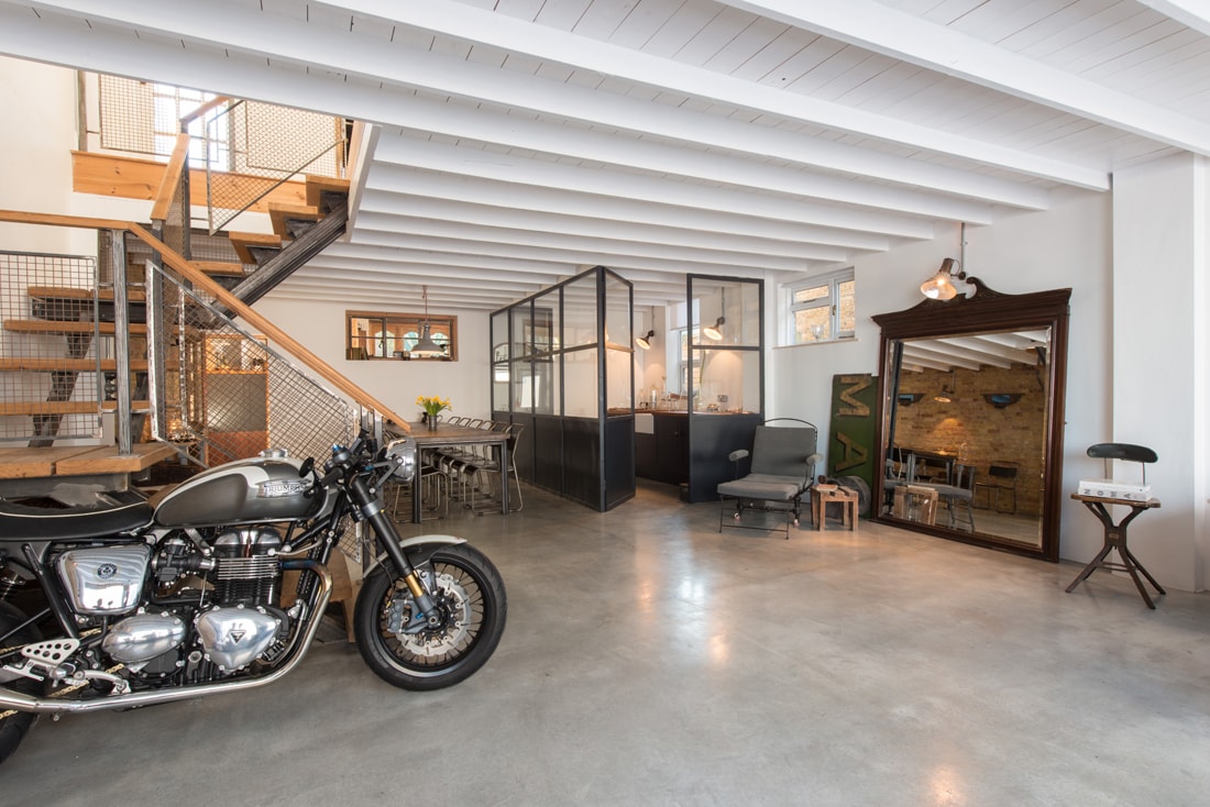 An open-concept living area showcases polished concrete flooring and sleek white beams overhead. A motorcycle is displayed near a staircase, which leads to additional spaces. A dining table is situated in the background, alongside a stylish glass partition and a vintage mirror.