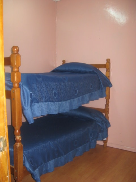 A compact room features a wooden bunk bed covered with blue bed linens. The walls are painted a soft pink, and the floor is a smooth wooden surface, creating a simple yet comfortable sleeping space.