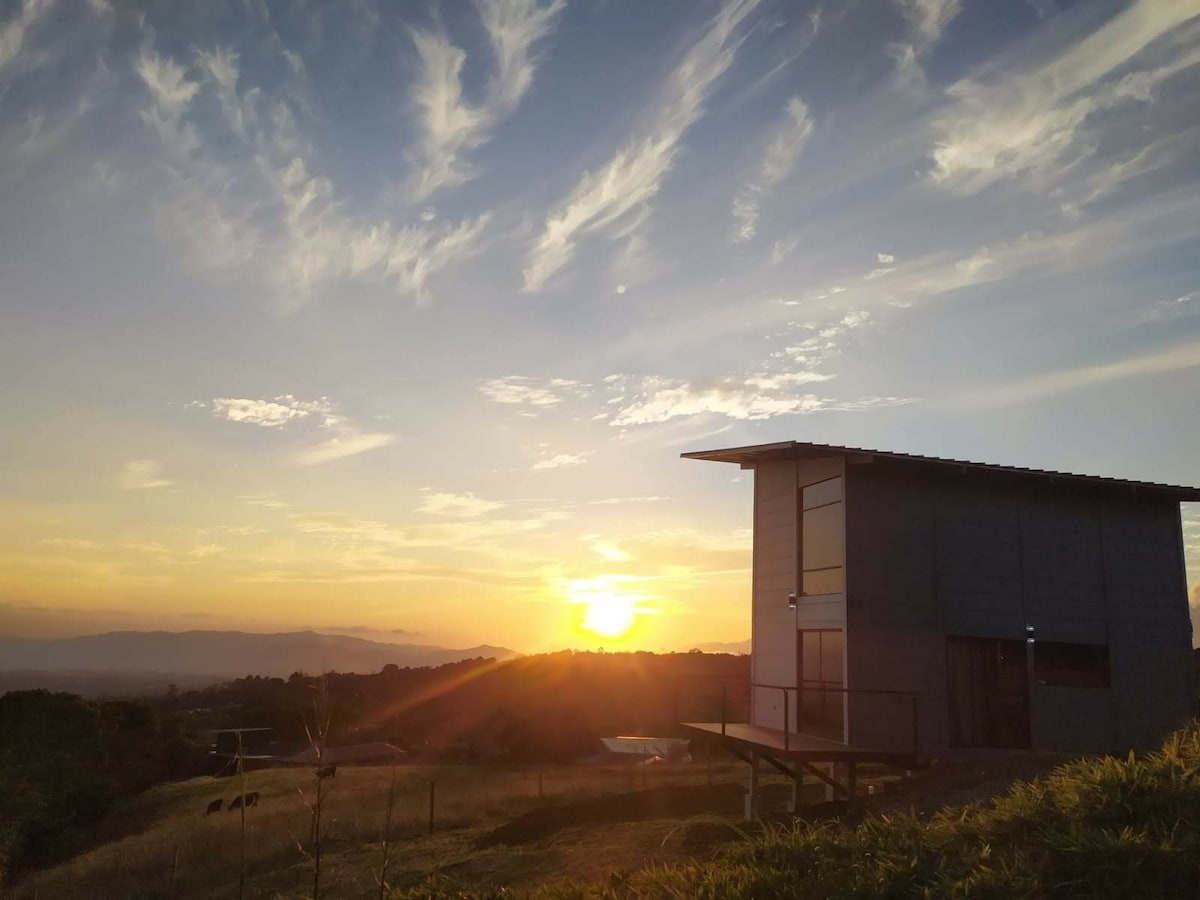 A modern loft is positioned on a hillside, with a stunning view of a sunset casting warm colors across the sky. The structure features large windows and a slanted roof, surrounded by lush green fields and scenic hills in the background.
