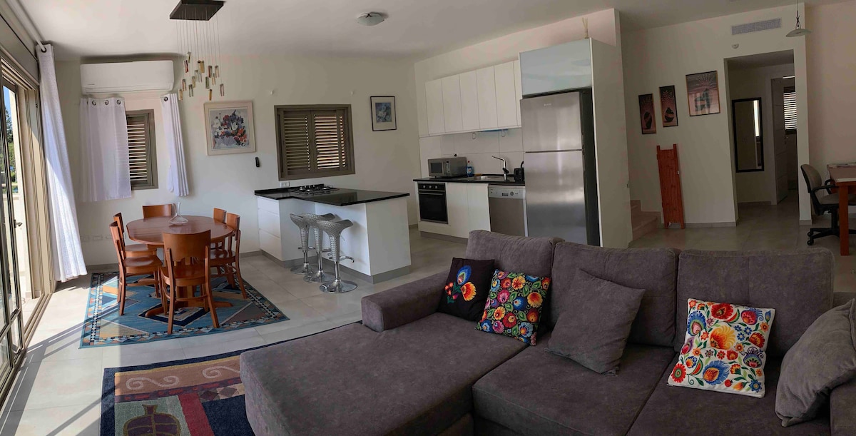 An open living space showcases a comfortable sectional sofa adorned with colorful cushions. A dining area with a round table for four sits adjacent to a modern kitchen equipped with stainless steel appliances. Natural light fills the room through large windows, creating an airy atmosphere.