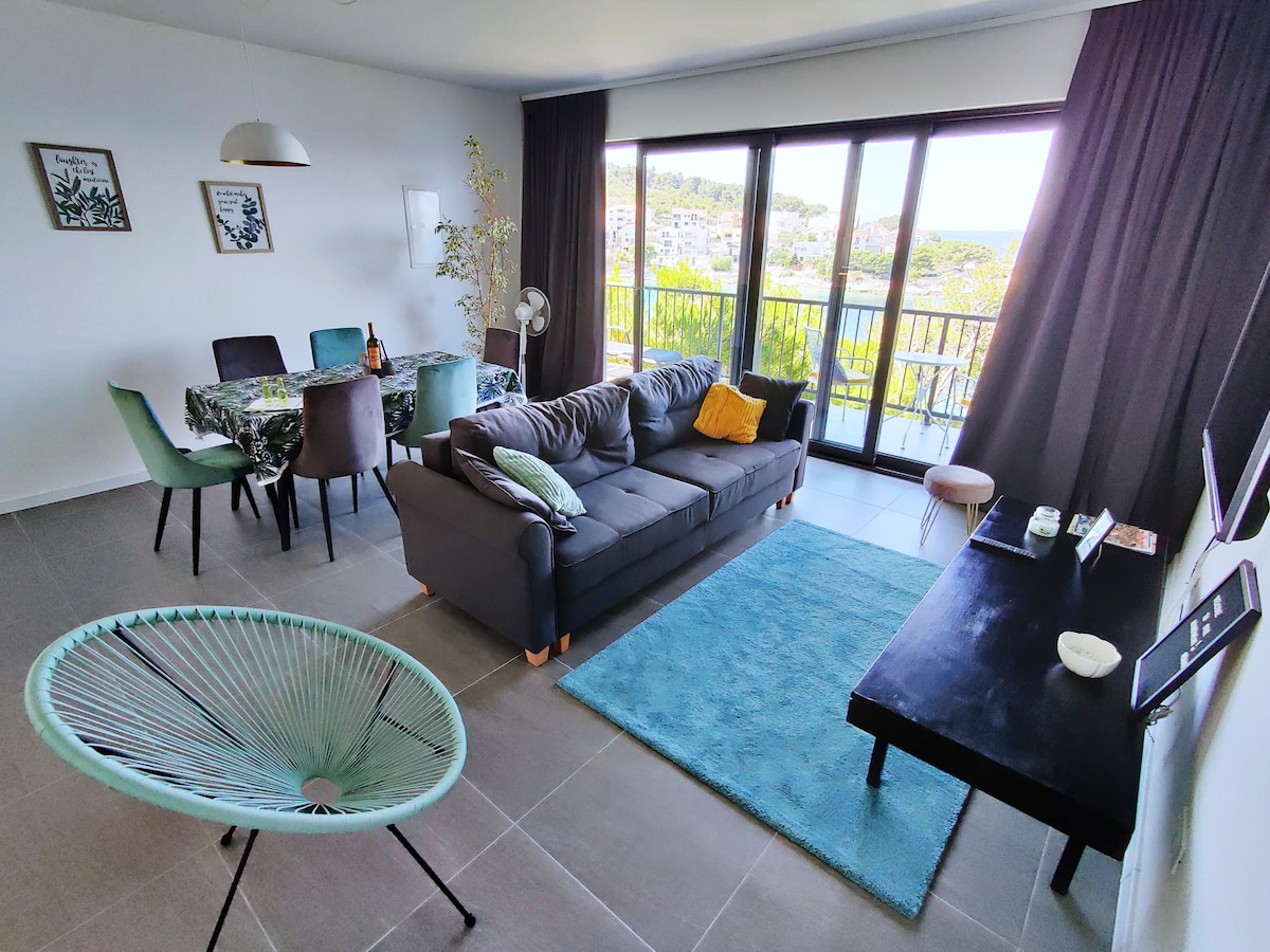 A bright living area features a spacious grey sofa with decorative cushions, a cozy teal rug, and a coffee table. A dining table with patterned tablecloth is surrounded by dining chairs. Expansive glass doors open to a balcony with scenic views, enhancing the airy atmosphere.