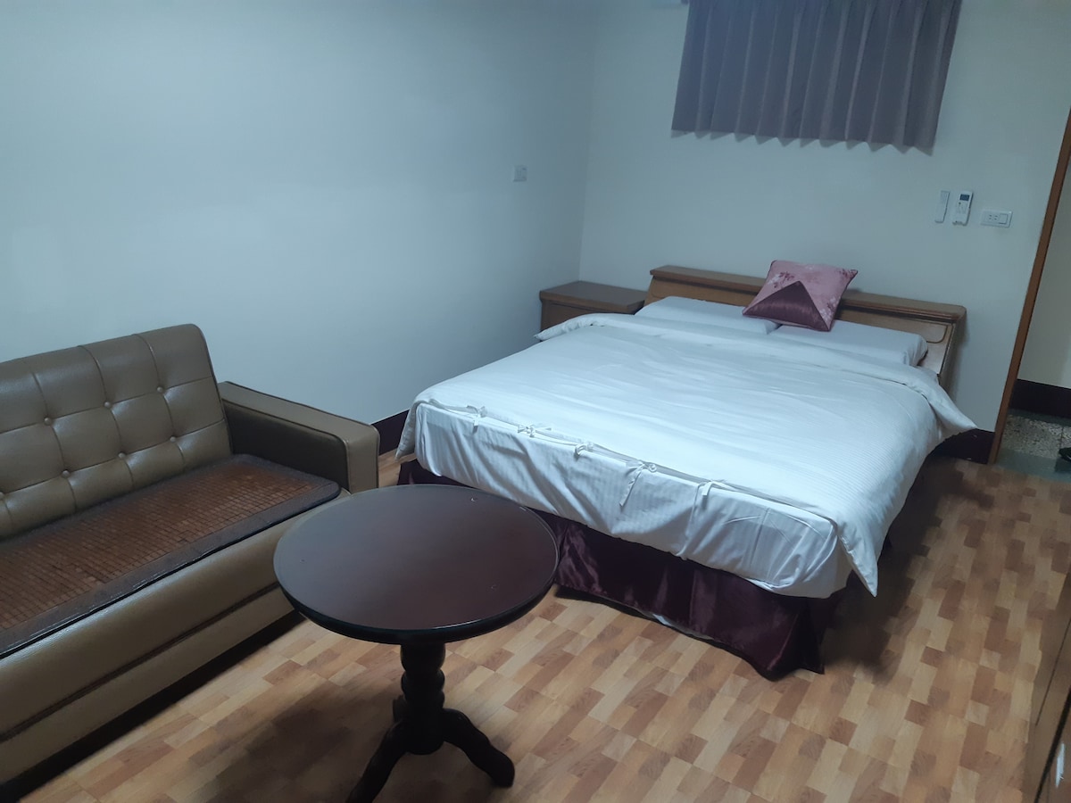 A well-arranged room features a spacious bed covered with a white linen and a decorative pillow. A brown sofa sits beside a small round table, while light flows in through a window adorned with sheer curtains.