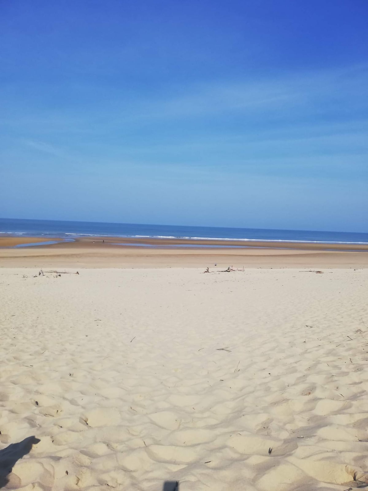 A sandy beach stretches towards the horizon, showcasing gentle waves lapping at the shore. The sky is a clear blue, blending seamlessly with the ocean. The expanse of sand leads to brief patches of water, inviting a serene and peaceful atmosphere.