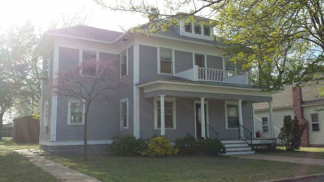 A three-story house is positioned under natural sunlight, highlighting its welcoming gray exterior. A covered porch with white railings extends from the front, flanked by trees and green foliage. A well-maintained lawn surrounds the base, contributing to a serene atmosphere.