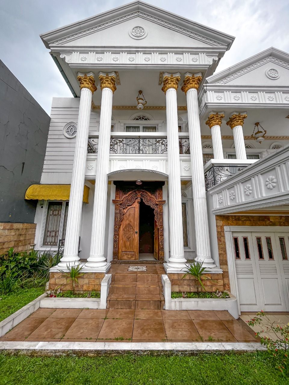 The entrance features grand white columns and an intricately carved wooden door, leading into the spacious residence. A tiled walkway is bordered by well-maintained greenery, providing a welcoming approach. The upper level showcases a balcony with decorative railings.