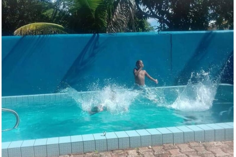 A private inground swimming pool is showcased, surrounded by a vibrant turquoise wall and tropical palm trees. Water splashes are seen as two individuals enjoy the refreshing space, highlighting a playful atmosphere in a sunlit outdoor setting.
