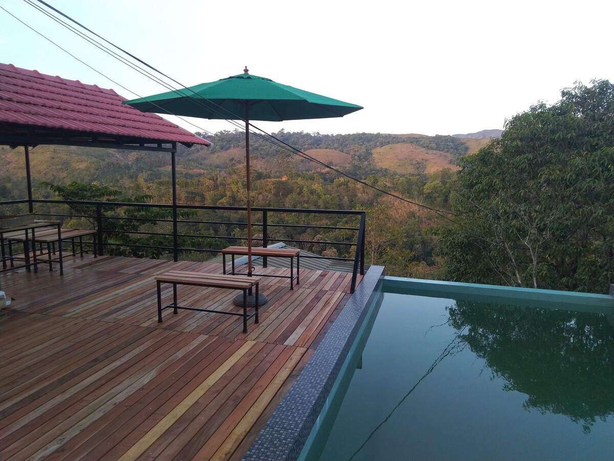 An outdoor deck features a tranquil infinity pool overlooking lush hills. A green umbrella provides shade over a wooden seating area, while the surrounding landscape showcases a blend of greenery and distant hills under a clear sky.