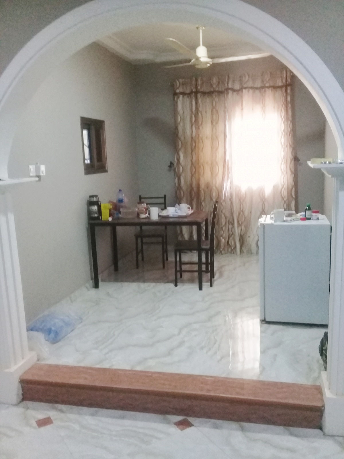 A dining area is visible through an arched entryway, featuring a wooden table surrounded by four chairs. A small refrigerator stands nearby, while natural light filters through patterned curtains that frame a window. The floor is covered in glossy tiles.