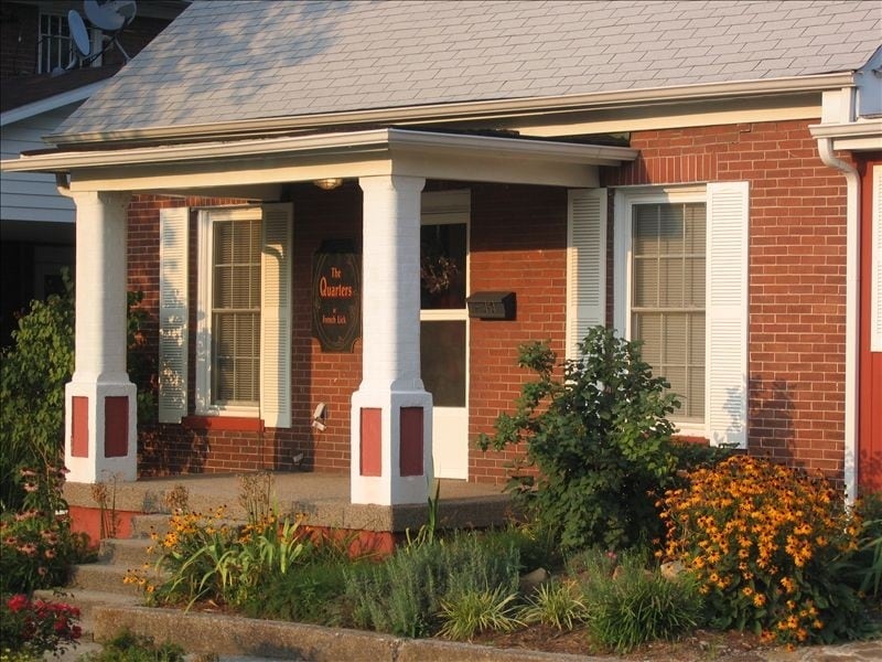 The exterior of a charming red brick cottage is displayed, featuring white columns and large windows. A welcoming porch is adorned with colorful flowers and greenery, adding to the inviting appearance of the home. The roof is gently sloped, complementing the cottage's classic design.