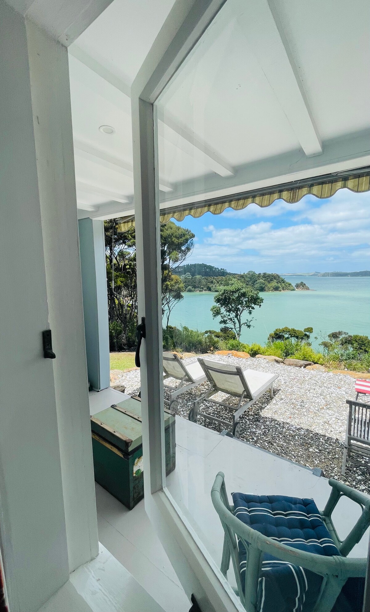 A view through a glass door frames a patio area with recliners facing a serene bay. Lush greenery and sandy shores are visible, enhancing the outdoor ambiance. The scene is bathed in natural light, inviting a sense of relaxation and connection with nature.
