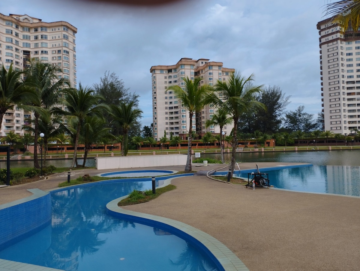 A serene pool area is surrounded by palm trees, with gentle curves leading to still waters. Lush greenery is visible along the water's edge, while modern buildings rise in the background, creating a tranquil environment for relaxation.