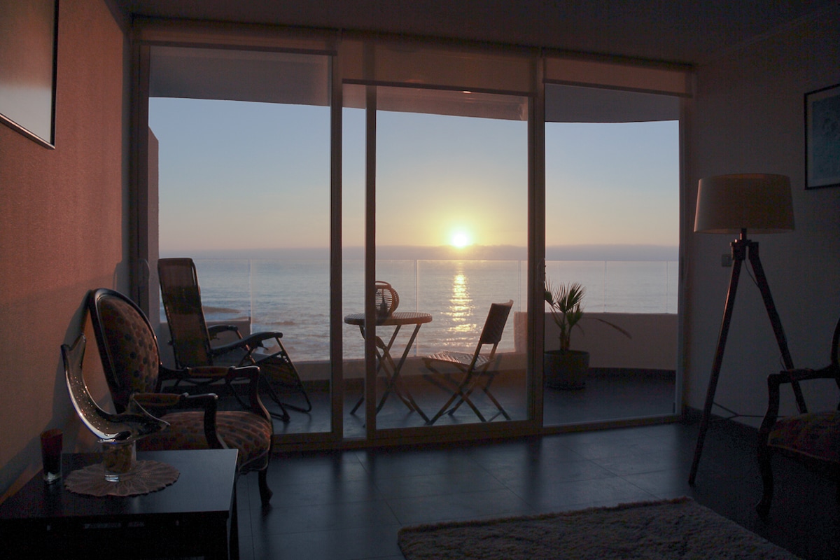 A serene view from the living space shows the sun setting over the calm sea, with a glass balcony that offers a clear perspective. Outdoor furniture, including a small table and chairs, is positioned for enjoying the tranquil surroundings. Warm light fills the room, enhancing the relaxed atmosphere.
