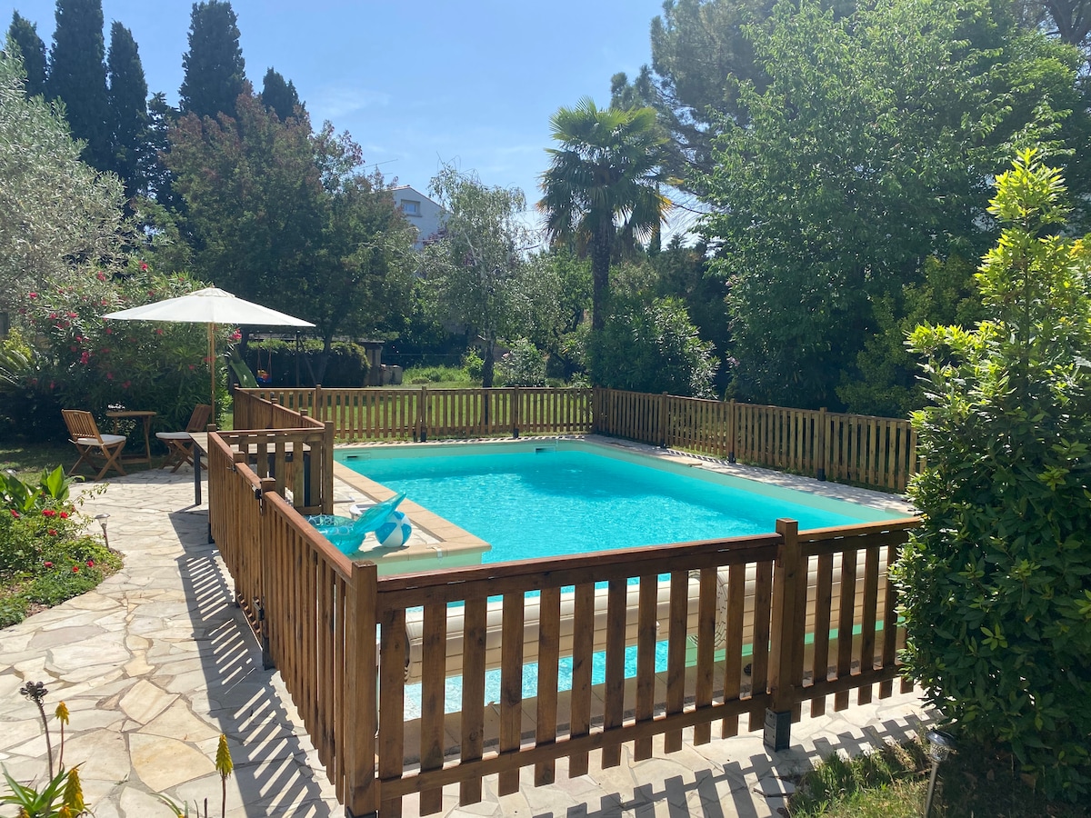 A tranquil pool area is enclosed by a wooden fence, featuring a refreshing water surface under dappled sunlight. A shaded lounge area with an umbrella and two chairs sits nearby, surrounded by lush greenery and tropical plants.