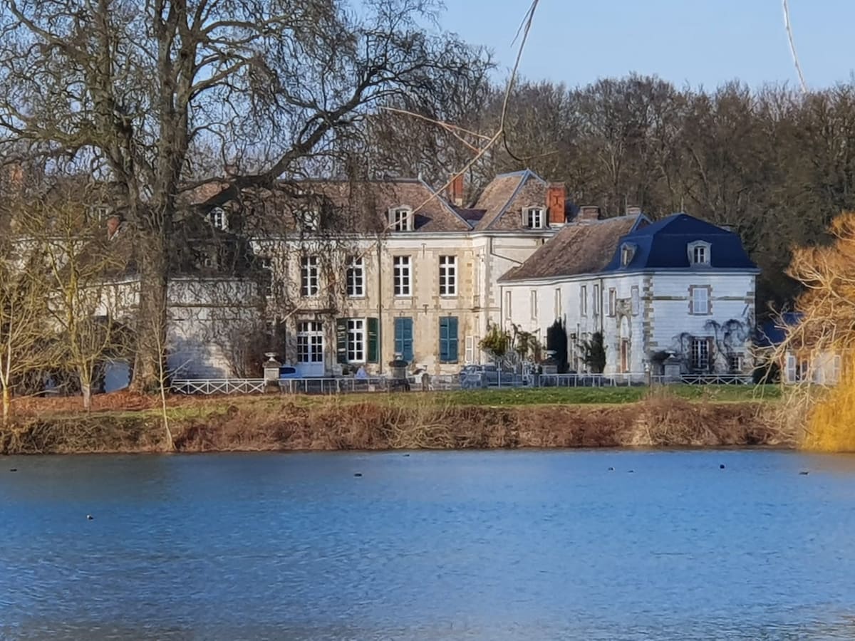 A historic chateau is viewed from across a tranquil lake, featuring a combination of stone facades with large windows and blue shutters. A serene landscape is visible with lush greenery surrounding the property, providing a peaceful setting.