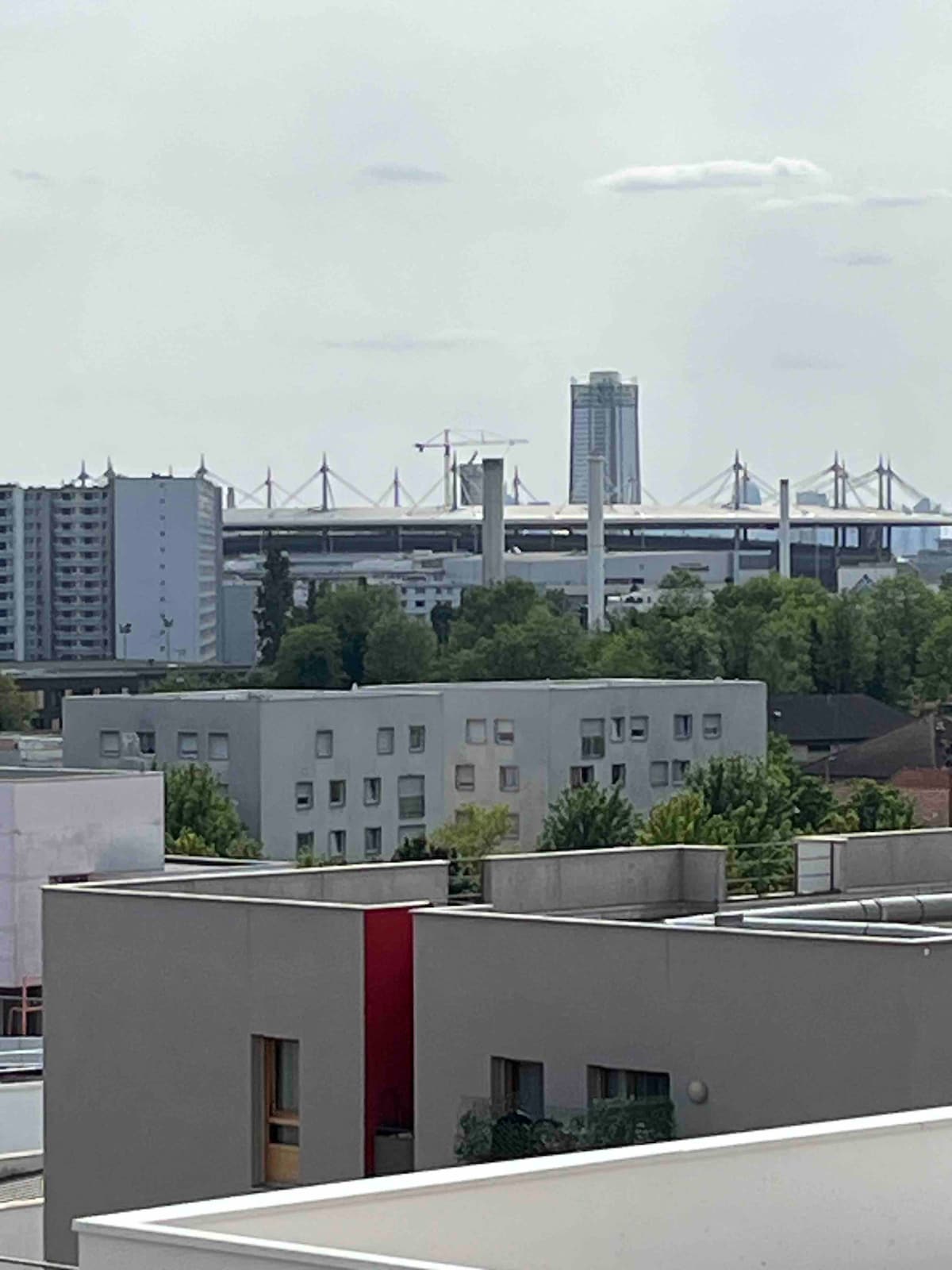 A cityscape view is captured from a rooftop, showcasing a blend of residential buildings with flat roofs and a modern architectural structure in the background. A large bridge and a tall building are visible, surrounded by greenery and under a cloudy sky.