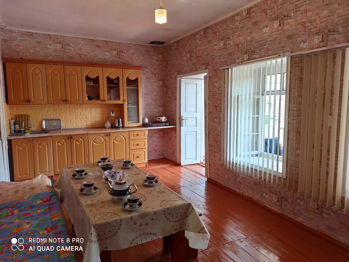 A dining area features a table set for multiple guests, complete with decorative tableware. Light-colored cabinetry provides storage in the adjacent kitchen, which is complemented by natural light from a window. The walls are adorned with rustic wallpaper, enhancing the warm ambiance.