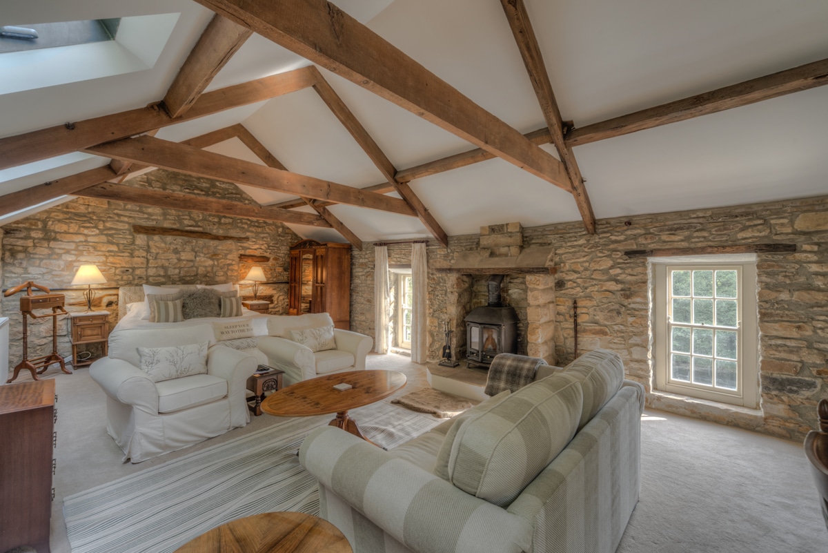 A spacious living area features exposed wooden beams and stone walls, complemented by cozy furnishings. A comfortable sofa and armchairs are arranged around a wooden coffee table, with soft light streaming through windows. A rustic fireplace adds to the inviting atmosphere, enhancing the room's charm.