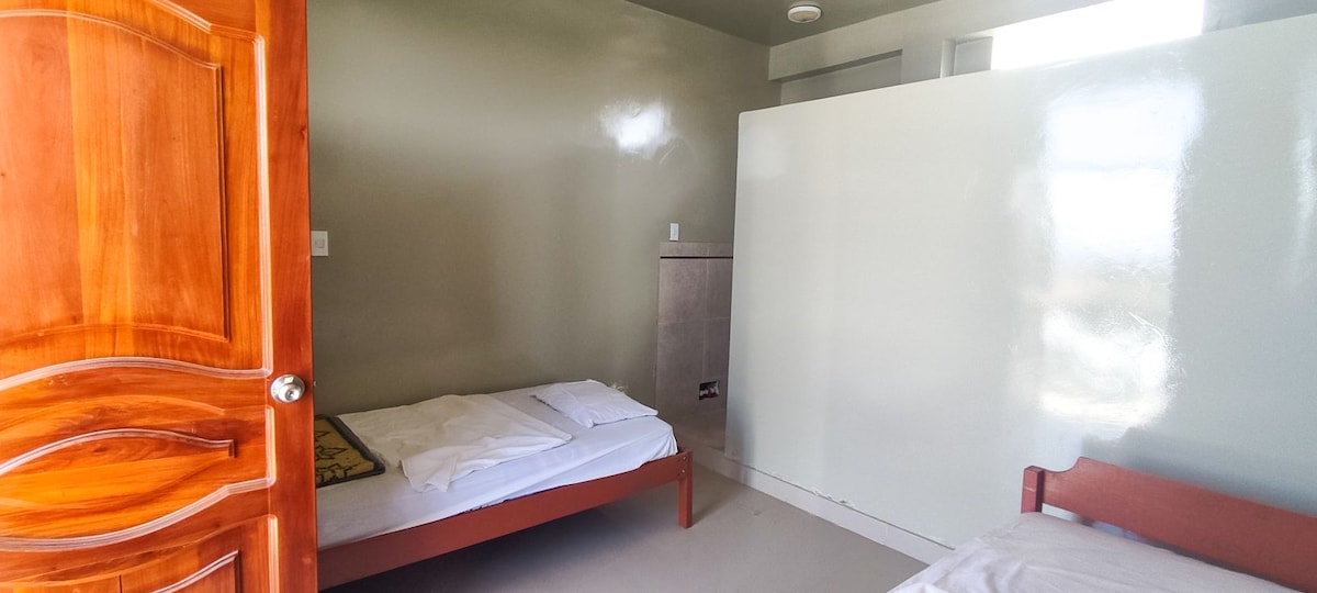 A simple guest room features two beds with clean linens. A solid wall divides the space, and a wooden door provides entry. Natural light enters through a window in the corner, illuminating the light green walls and neutral flooring.