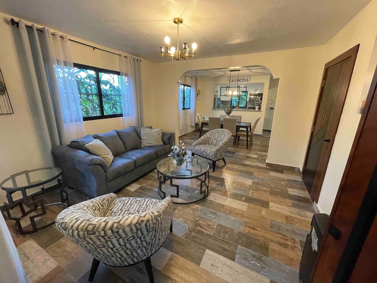 A bright living area features a gray sofa with two patterned armchairs, arranged around a small table. A large window allows natural light to fill the space, while a dining area can be seen in the background, separated by an archway.