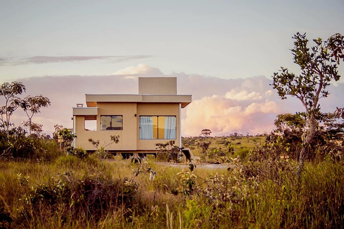A spacious bungalow is situated in an open natural landscape, surrounded by greenery. Large windows reflect the soft colors of the sky during dusk, while distant hills can be seen in the background. The peaceful environment showcases the serene setting of the Chapada dos Veadeiros.
