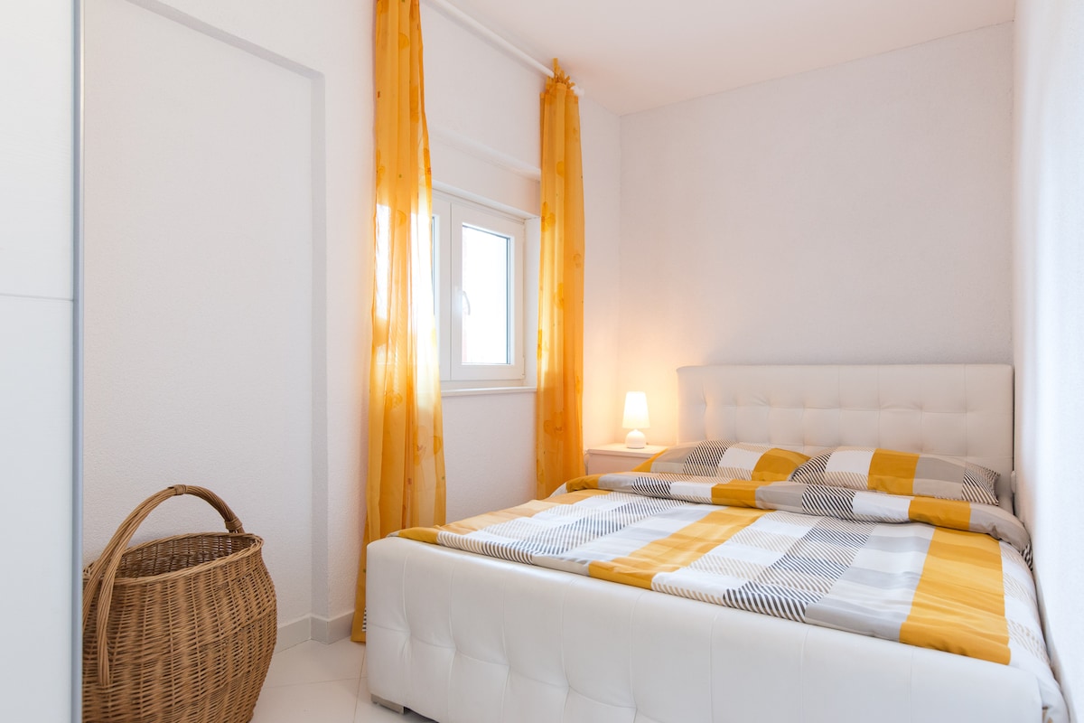 A bright bedroom features a spacious bed dressed in yellow and gray bedding. Soft curtains frame the window, allowing natural light to illuminate the room. A bedside lamp is visible, complemented by a wicker basket positioned nearby.